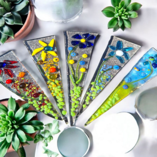 An overhead view of four artisanal fused glass plant stakes, triangular in shape. Each stake features vibrant, raised floral and botanical elements in various colors like yellow, blue, and green, suggesting the cottagecore aesthetic. The textured clear glass background of the stakes highlights the intricate designs, positioned next to three succulent plants.