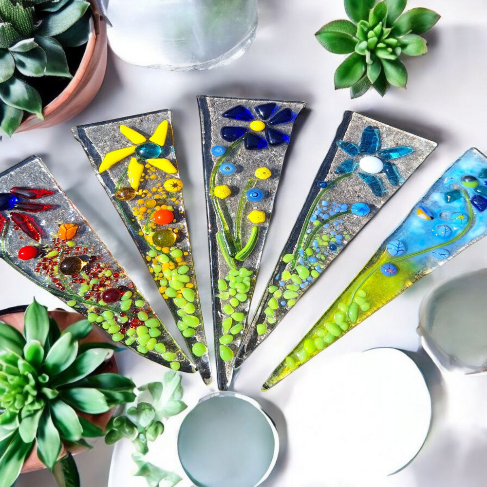 An overhead view of four artisanal fused glass plant stakes, triangular in shape. Each stake features vibrant, raised floral and botanical elements in various colors like yellow, blue, and green, suggesting the cottagecore aesthetic. The textured clear glass background of the stakes highlights the intricate designs, positioned next to three succulent plants.