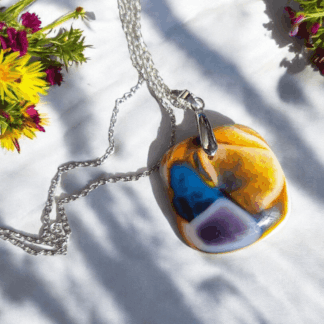 Handmade fused glass pendant necklace with swirls of yellow, blue, and purple, hanging from a silver-plated chain, displayed on white fabric with colourful flowers.