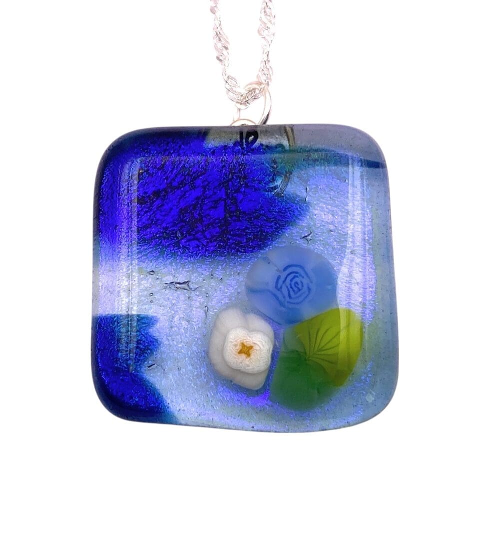 A vibrant blue glass pendant featuring floral designs, including a white flower and green leaves, suspended by a silver chain.
