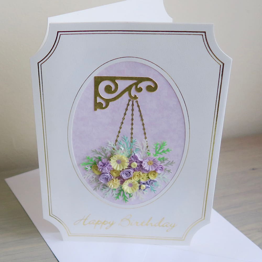 A handmade birthday card featuring a hanging basket filled with lilac and pink quilled flowers with a Happy Birthday message in gold foil