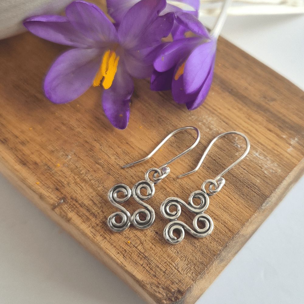 Overhead shot of double-ended spiral earrings lying on a piece of wood with purple flowers nearby.