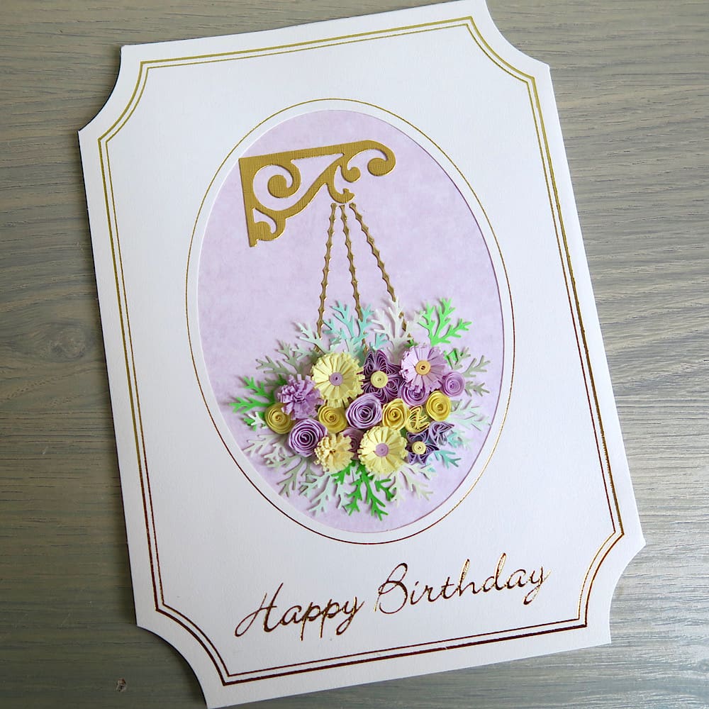 A handmade birthday card featuring a hanging basket filled with lilac and pink quilled flowers with a Happy Birthday message in gold foil