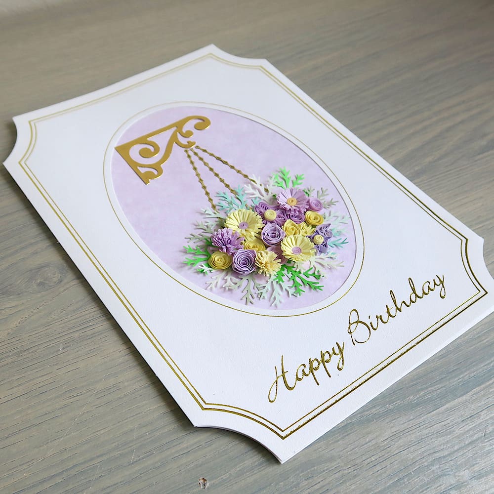 A handmade birthday card featuring a hanging basket filled with lilac and pink quilled flowers with a Happy Birthday message in gold foil