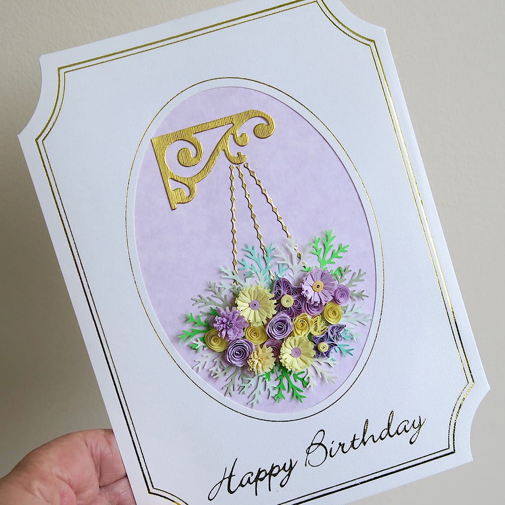 A handmade birthday card featuring a hanging basket filled with lilac and pink quilled flowers with a Happy Birthday message in gold foil