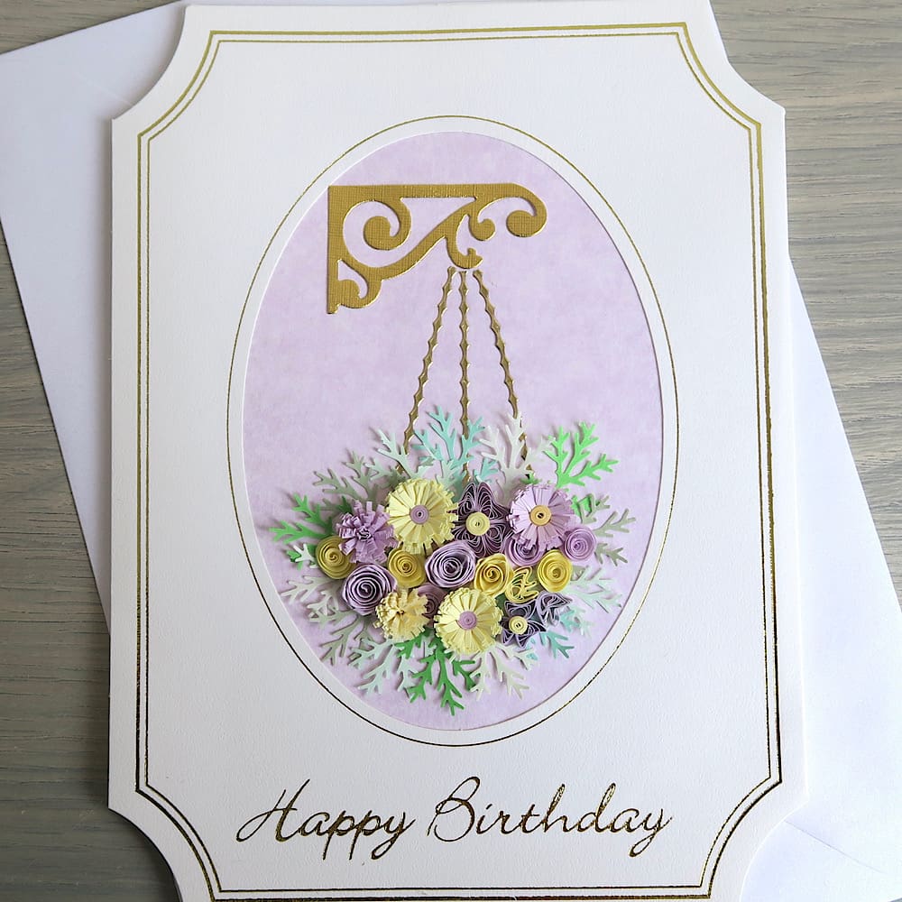 A handmade birthday card featuring a hanging basket filled with lilac and pink quilled flowers with a Happy Birthday message in gold foil