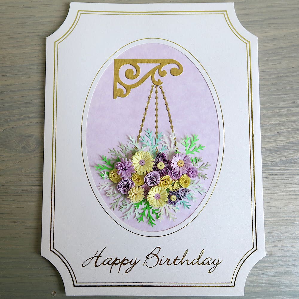 A handmade birthday card featuring a hanging basket filled with lilac and pink quilled flowers with a Happy Birthday message in gold foil