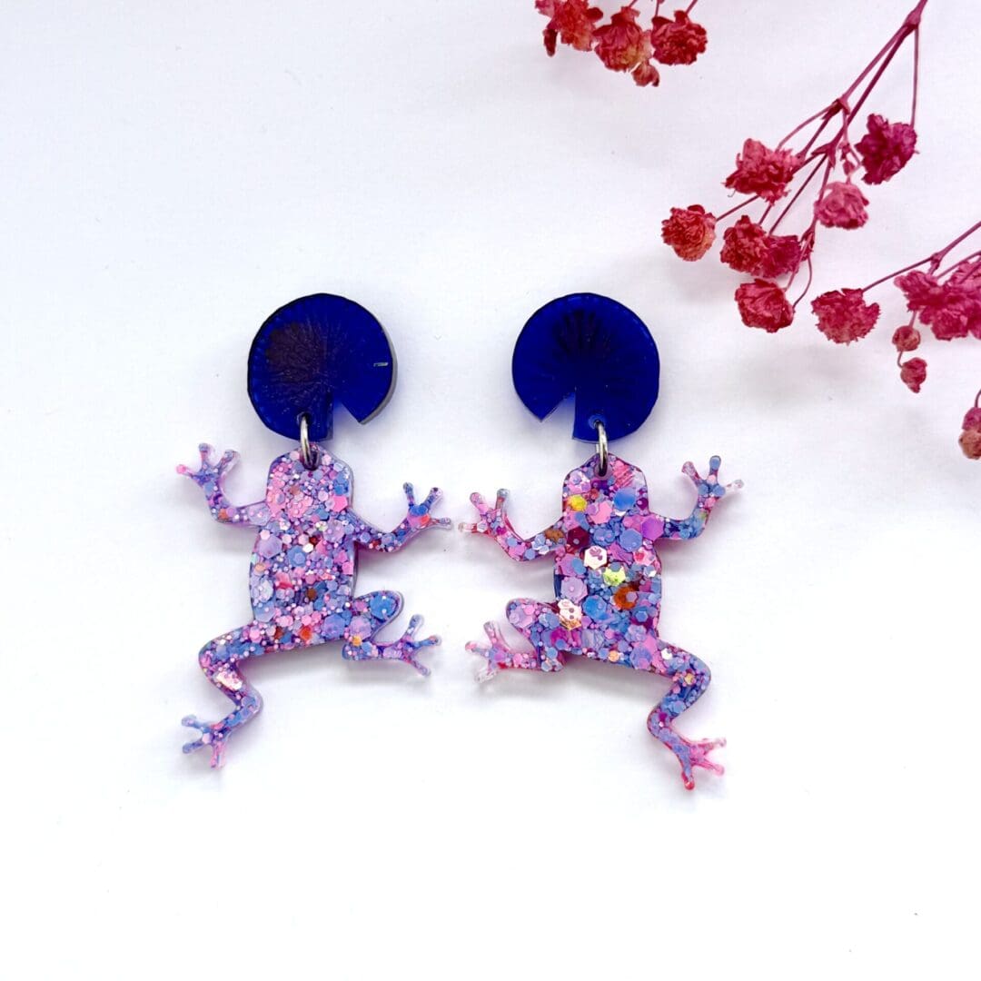 Cute pink and purple glitter climbing frog resin earrings hanging from blue lily pad studs.