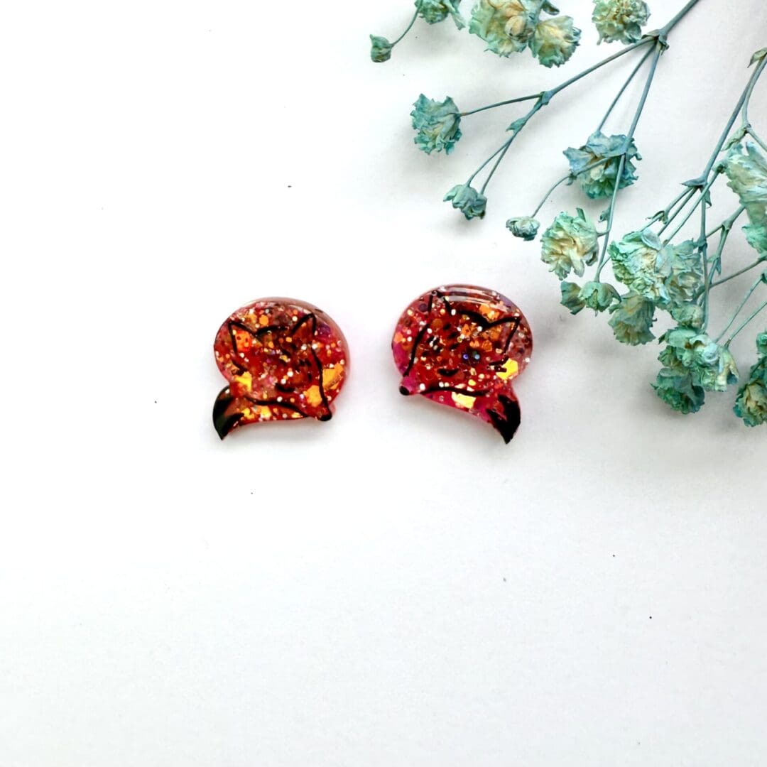 A pair of copper and orange glitter sleeping fox surgical steel studs earrings, each facing a different direction and curled up. Image painted with black to show line work.
