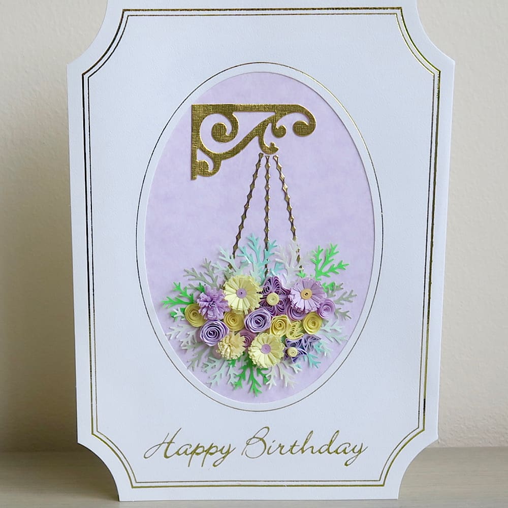 A handmade birthday card featuring a hanging basket filled with lilac and pink quilled flowers with a Happy Birthday message in gold foil