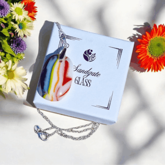 A handmade glass pendant with vibrant stripes of red, orange, yellow, green, blue, and white hangs from a silver chain next to its white gift box with "Sandgate Glass" written on it. To the left, a bouquet of purple, white, and yellow flowers is visible, and on the right, an orange flower.