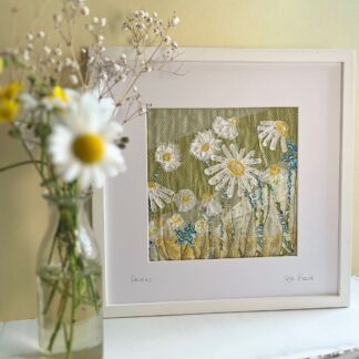Square framed textile art of a field of yellow and white daisies on a green background with a vase of daisies in front