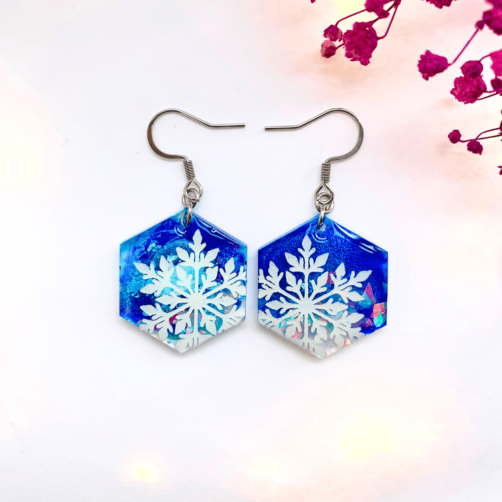 Pair of hexagon-shaped resin earrings with vibrant blue and iridescent backgrounds, each featuring a detailed white snowflake design. Earrings are photographed against a white background with pink dried flowers