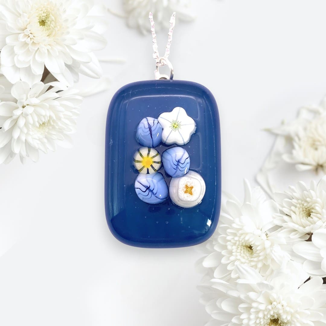 A blue handmade glass pendant featuring intricate three dimensional floral designs in white and blue against a soft backdrop of white flowers.