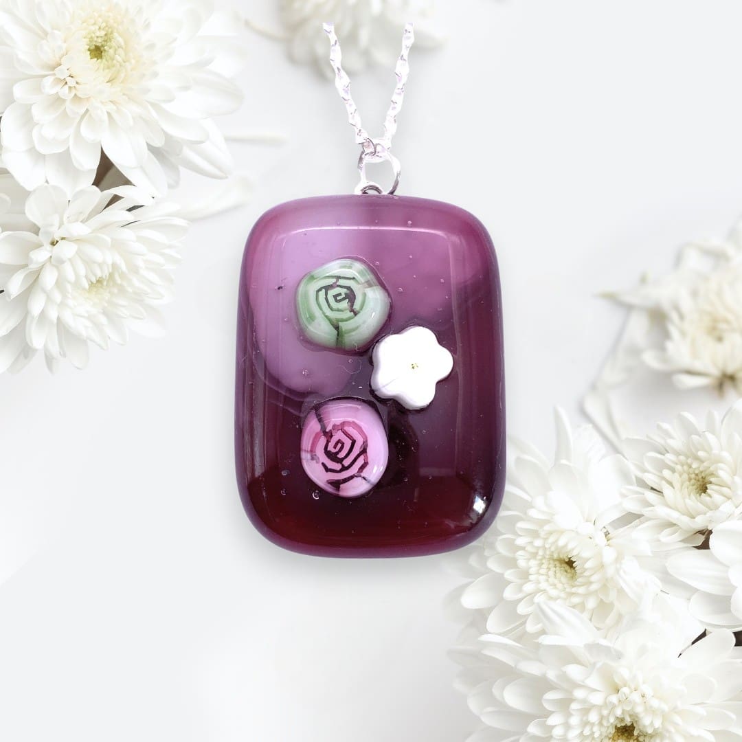 A fuchsia coloured handmade glass pendant adorned with three dimensional pink and green rose designs and a white flower, resting among white flowers.