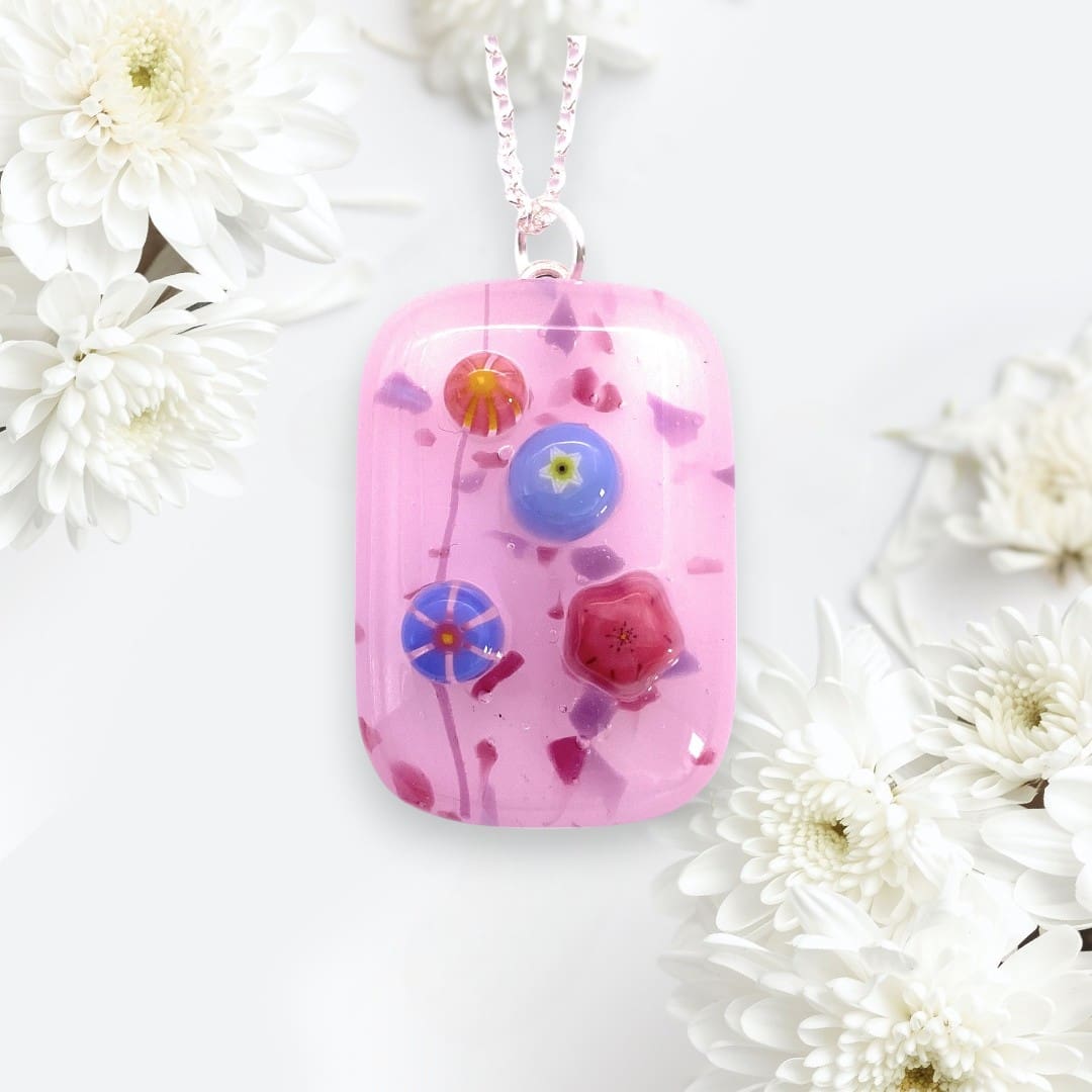 A pink handmade glass pendant adorned with colorful three dimensional floral designs, hanging on a delicate chain against a backdrop of white daisies.