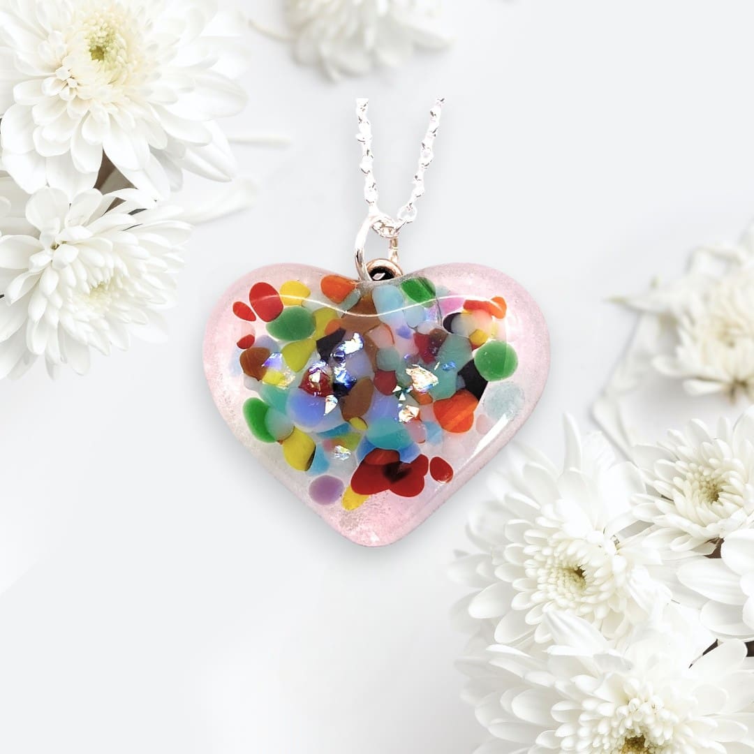 A handmade heart-shaped pendant made of dichroic glass. The pendant is filled with colorful, abstract patterns and flecks of shimmering material, creating a vibrant, eye-catching design. It hangs from a delicate silver chain. The background is soft and elegant, decorated with white chrysanthemum flowers, enhancing the pendant's delicate and artistic appearance.