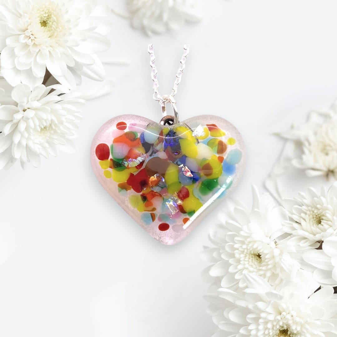 A handmade heart-shaped pendant made of dichroic glass. The pendant is filled with colorful, abstract patterns and flecks of shimmering material, creating a vibrant, eye-catching design. It hangs from a delicate silver chain. The background is soft and elegant, decorated with white chrysanthemum flowers, enhancing the pendant's delicate and artistic appearance.