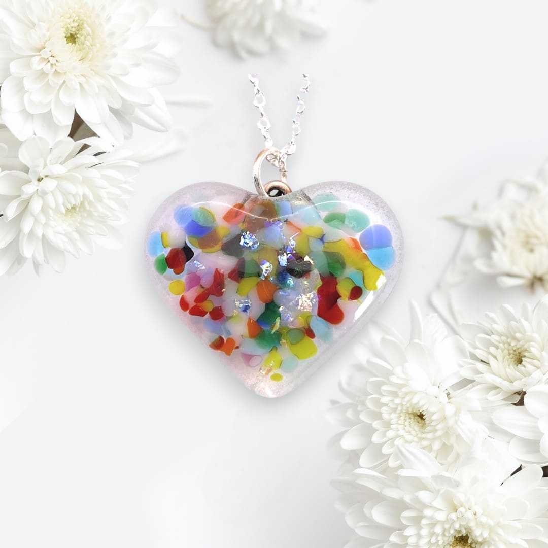 A handmade heart-shaped pendant made of dichroic glass. The pendant is filled with colorful, abstract patterns and flecks of shimmering material, creating a vibrant, eye-catching design. It hangs from a delicate silver chain. The background is soft and elegant, decorated with white chrysanthemum flowers, enhancing the pendant's delicate and artistic appearance.