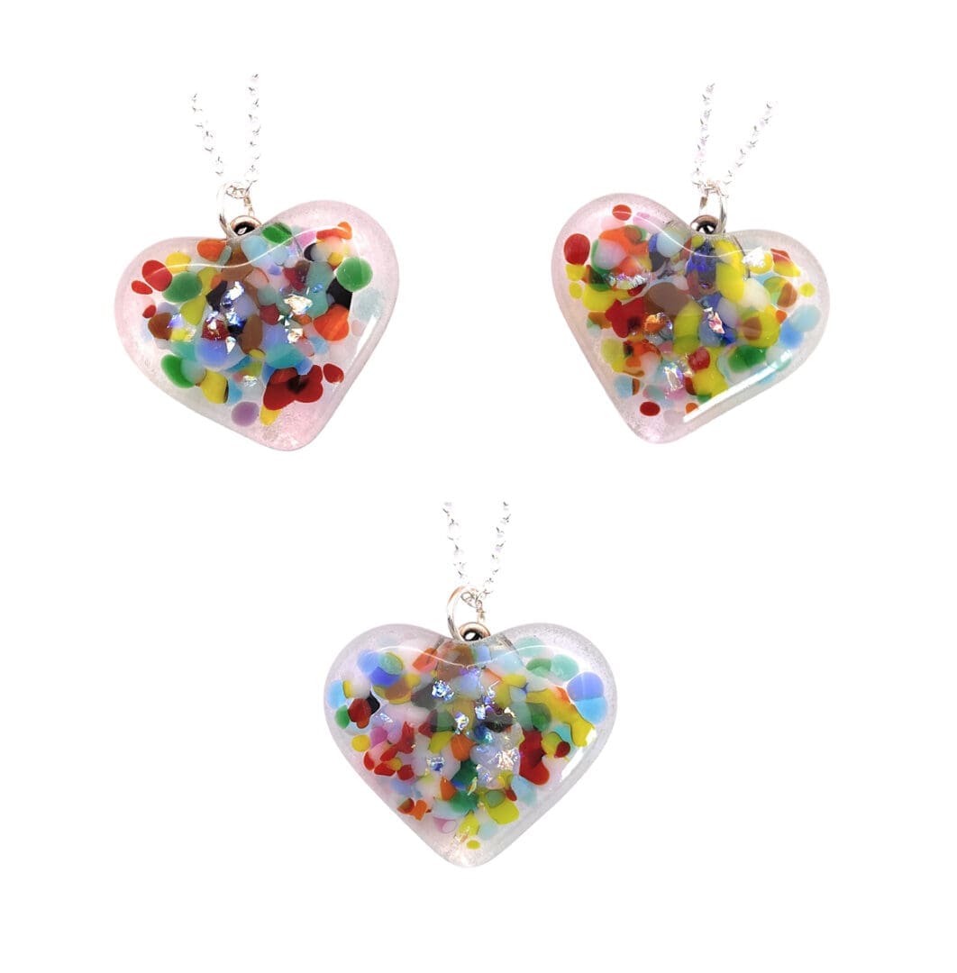 Three handmade heart-shaped pendants made of clear glass. Each pendant contains colorful, confetti-like pieces and shiny dichroic glass fragments embedded inside, creating a vibrant and eye-catching design. The pendants are suspended from delicate silver chains.