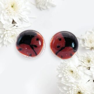 Close-up of a pair of handmade glass red and black ladybird earrings resting among white chrysanthemums on a light background.