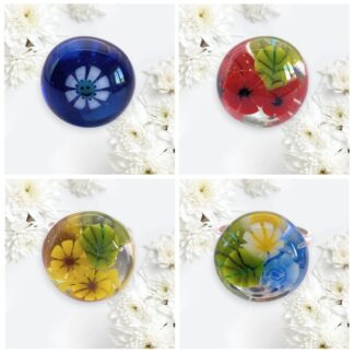 Four colourful handmade glass rings featuring embedded flowers on a white floral background, showcasing blue, red, yellow, and green designs.