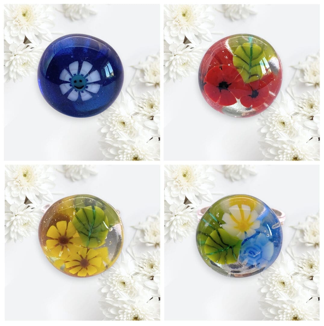 Four colourful handmade glass rings featuring embedded flowers on a white floral background, showcasing blue, red, yellow, and green designs.