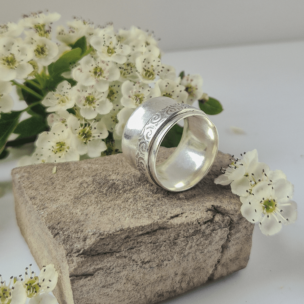 Handmade silver spinner ring resting on its side on a piece of rock, surrounded by white flowers.