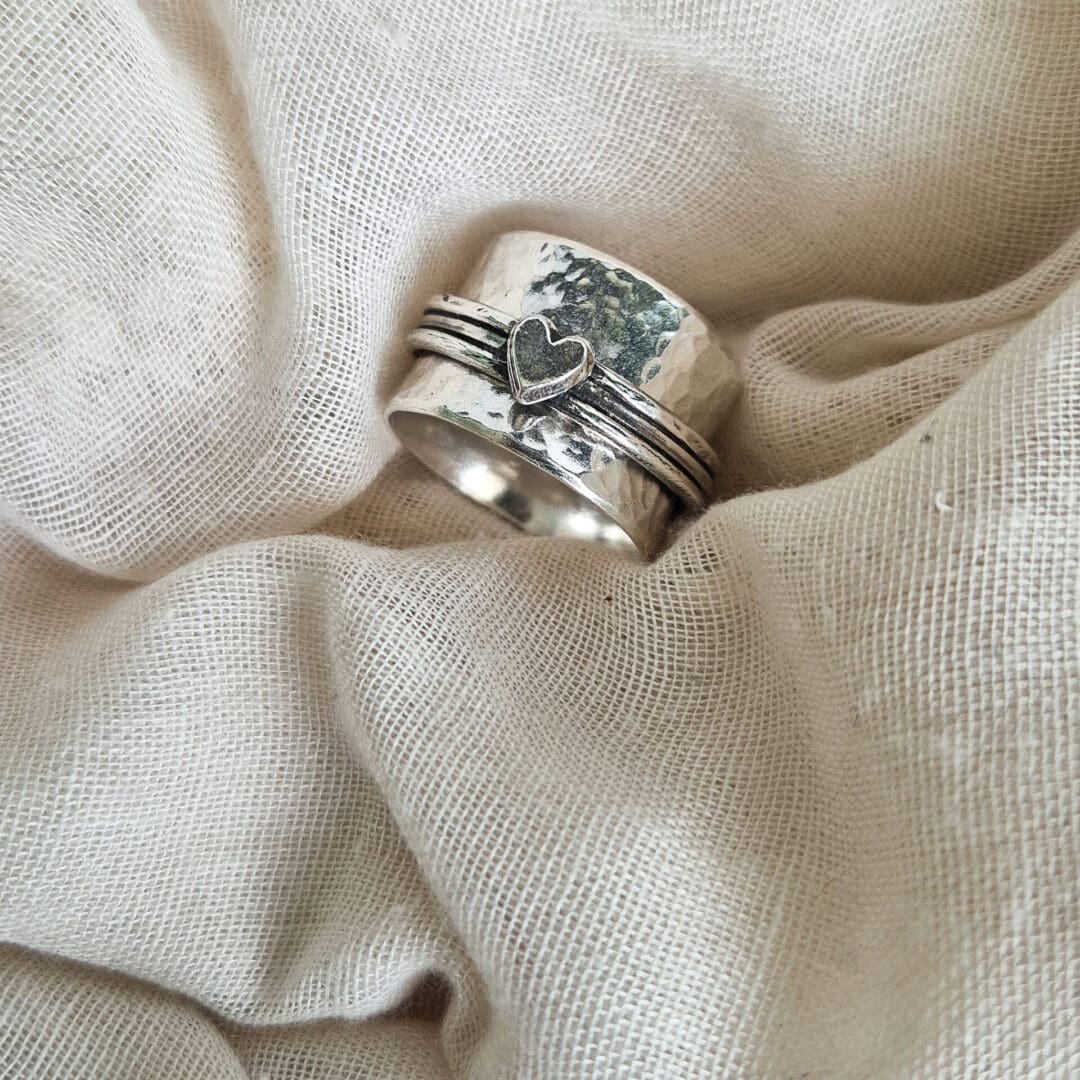 Chunky sterling silver spinner ring with hammered texture and heart embellishment against a white fabric background