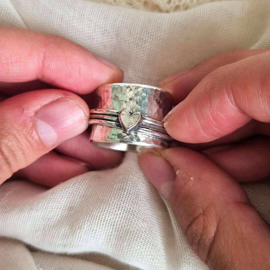 Chunky sterling silver spinner ring with hammered texture and heart embellishment being held against a white fabric background.