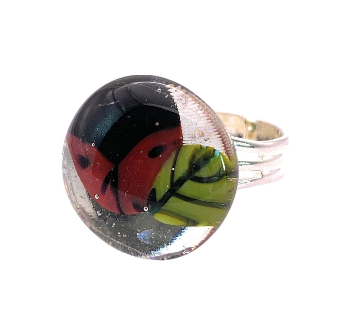 A stylish ring featuring a round glass cabochon with a vibrant red ladybird and green leaf, mounted on a shiny silver band.