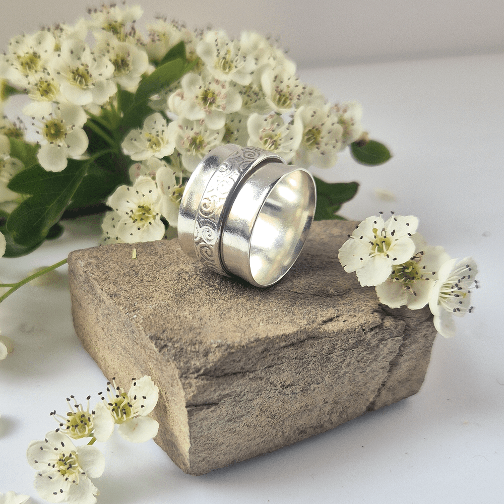 Handcrafted sterling silver spinner ring resting on its side on a piece of rock, surrounded by white flowers.