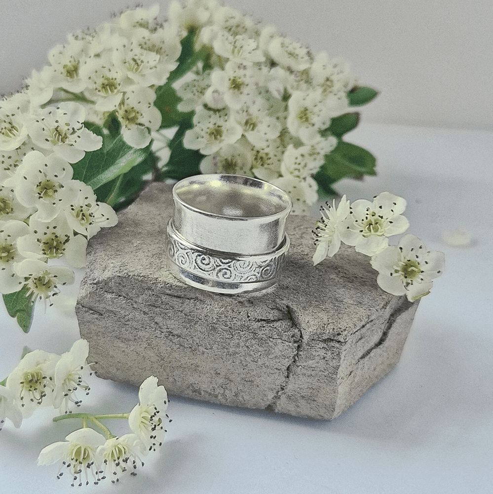 Handmade silver spinner ring resting on a piece of rock, surrounded by white flowers.