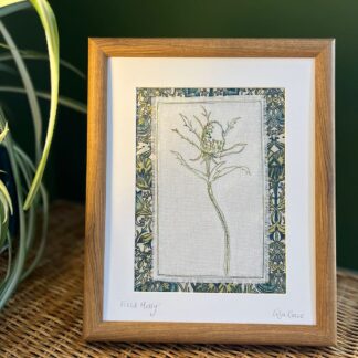 Framed textile art showing an embroidery of a single stem thistle on white linen fabric. Mounted on green floral fabric. Picture is standing on a table with a plant.