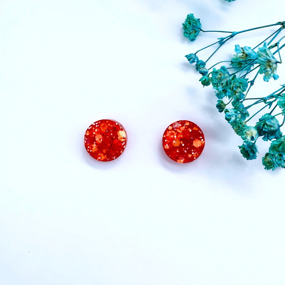 Handmade resin stud earrings in red and orange glitter, round shape with hypoallergenic backs, laid flat on a white background with blue dried flowers.