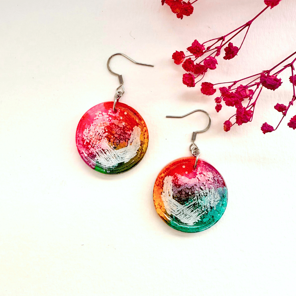A pair of round rainbow resin earrings inspired by petri dishes, with abstract swirls of pink, green, blue, orange, and white, placed beside pink dried flowers on a white background.