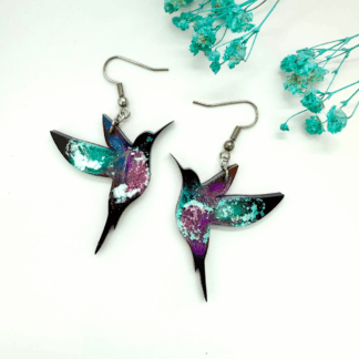 A pair of handmade resin hummingbird earrings in swirling galaxy tones of teal, purple and white, laid flat on a white background with blue dried flowers.