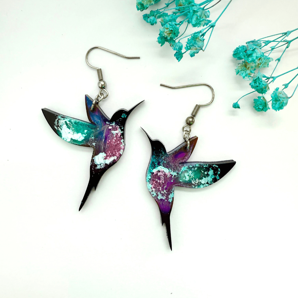 A pair of handmade resin hummingbird earrings in swirling galaxy tones of teal, purple and white, laid flat on a white background with blue dried flowers.