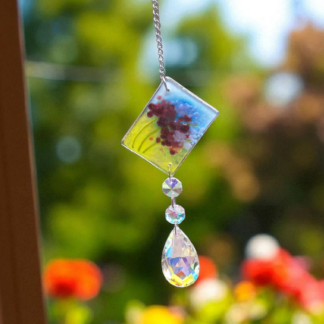 Handmade fused glass lavender suncatcher with teardrop crystal, hanging in a sunny window with a blurred flower garden in the background.
