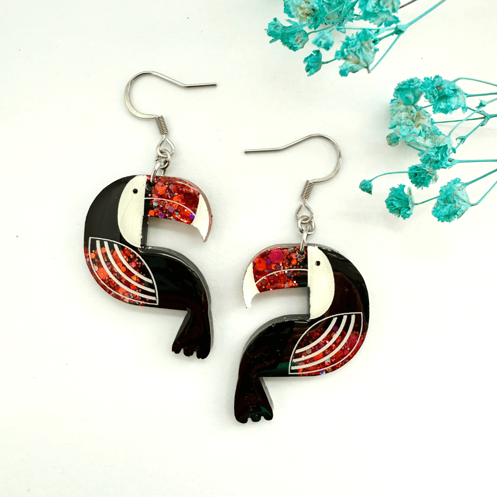 A pair of handmade resin toucan earrings with chunky red-orange glitter detailing, displayed on a white background.