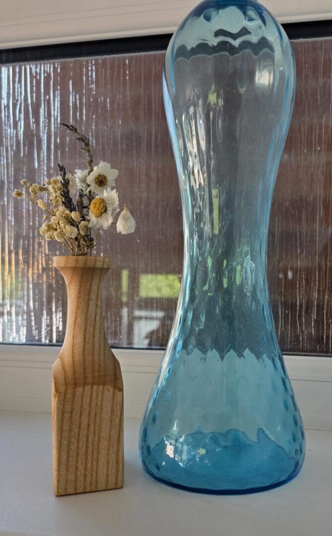 Handmade ash wood bud vase with a tall, elegant neck and square base, holding white and lavender dried flowers, displayed in front of a blue glass vase.