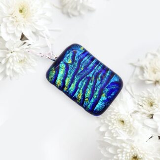 A vibrant handmade dichroic glass blue and green textured pendant hanging among white flowers, showcasing intricate patterns and a sleek design.