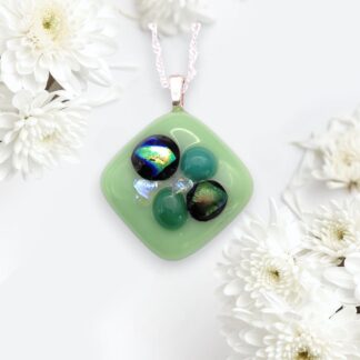 A square handmade glass green pendant adorned with iridescent black and turquoise stones, surrounded by white flowers.