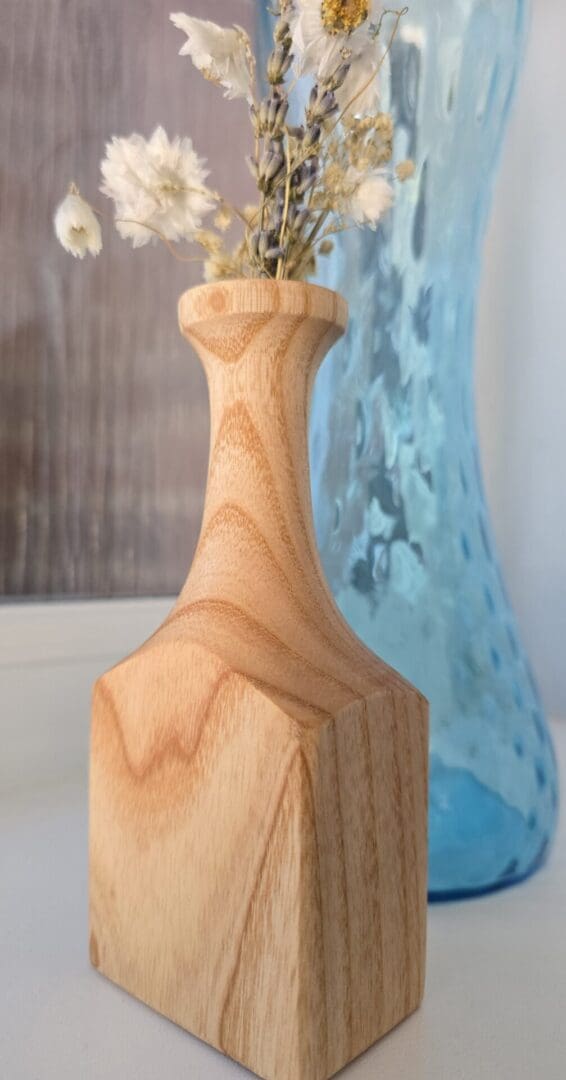 Handmade ash wood bud vase with a tall, elegant neck and square base, holding white and lavender dried flowers, displayed in front of a blue glass vase.