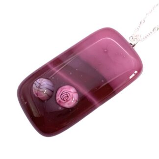 A rectangular pink pendant with two decorative millefiori glass pieces, a lilac leaf and a pink rose, suspended on a silver chain.
