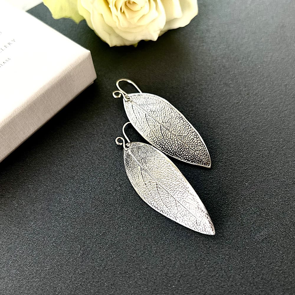 Fine silver clay sage leaf drop earrings with sterling silver ear wires. Displayed on a dark grey tile.