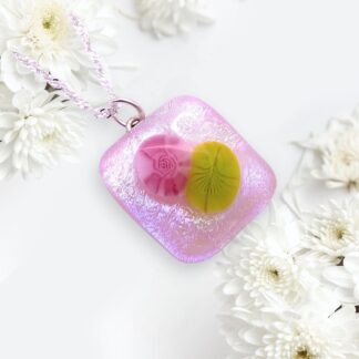 A vibrant handmade dichroic glass pendant in pink with floral designs, hanging from a silver chain, set against white daisies.