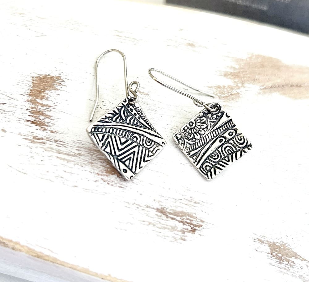 A pair of fine silver handcrafted square drop earrings that have a textured design with sterling silver ear wires. Displayed on white washed wood.