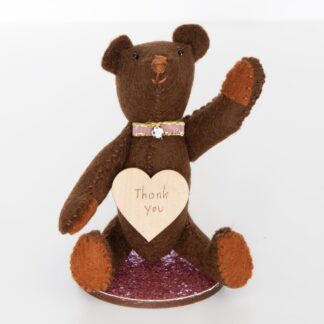 Dark brown felt bear with words, Thank You on a heart stuck to his chest and sitting on a pink glitter disk go wood.