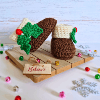 A pair of handmade chocolate brown crochet baby booties with a cream top and green holly leaf embellishments.
